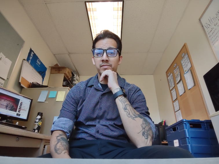 The image shows Alberto sitting in his office looking at the camera. He has dark short hair with glasses and a purple collared shirt. He also has tattoos on both arms. 