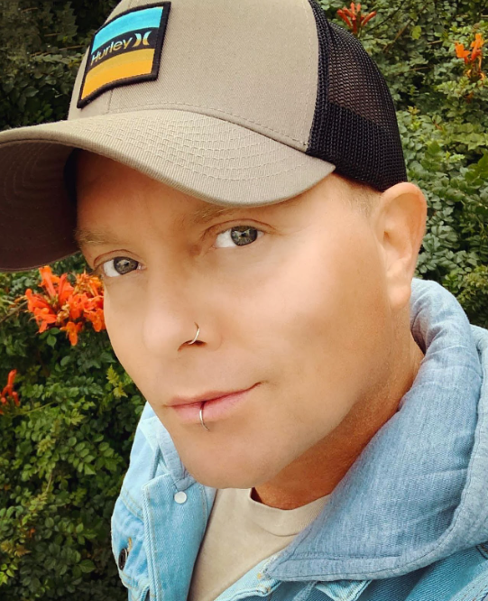 The image shows Brady Kerr outside smiling and looking at the camera. He has a hat on, and light hair and eyes. He has a lip ring and a nose ring. 