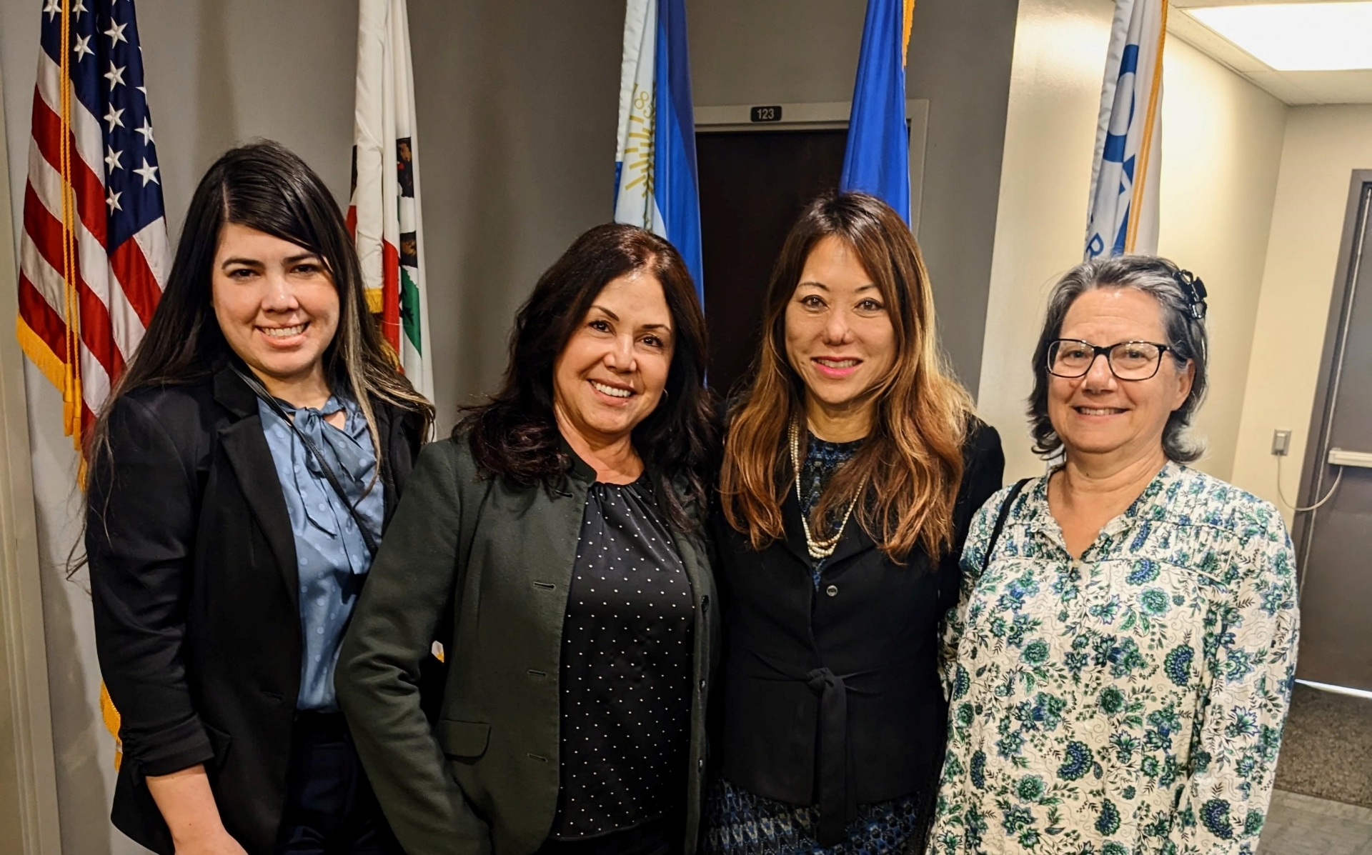 Four women stand smiling in front of a bank of flags.