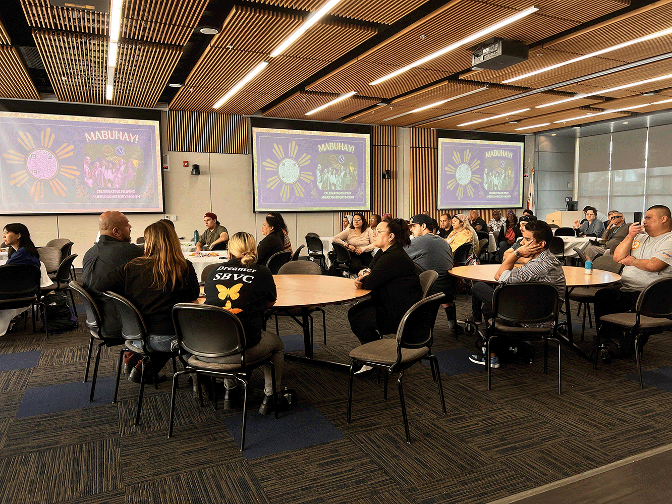 The image shows multiple groups of people sitting round round tables in a large room with multiple screens. The screens show the presentation for the event