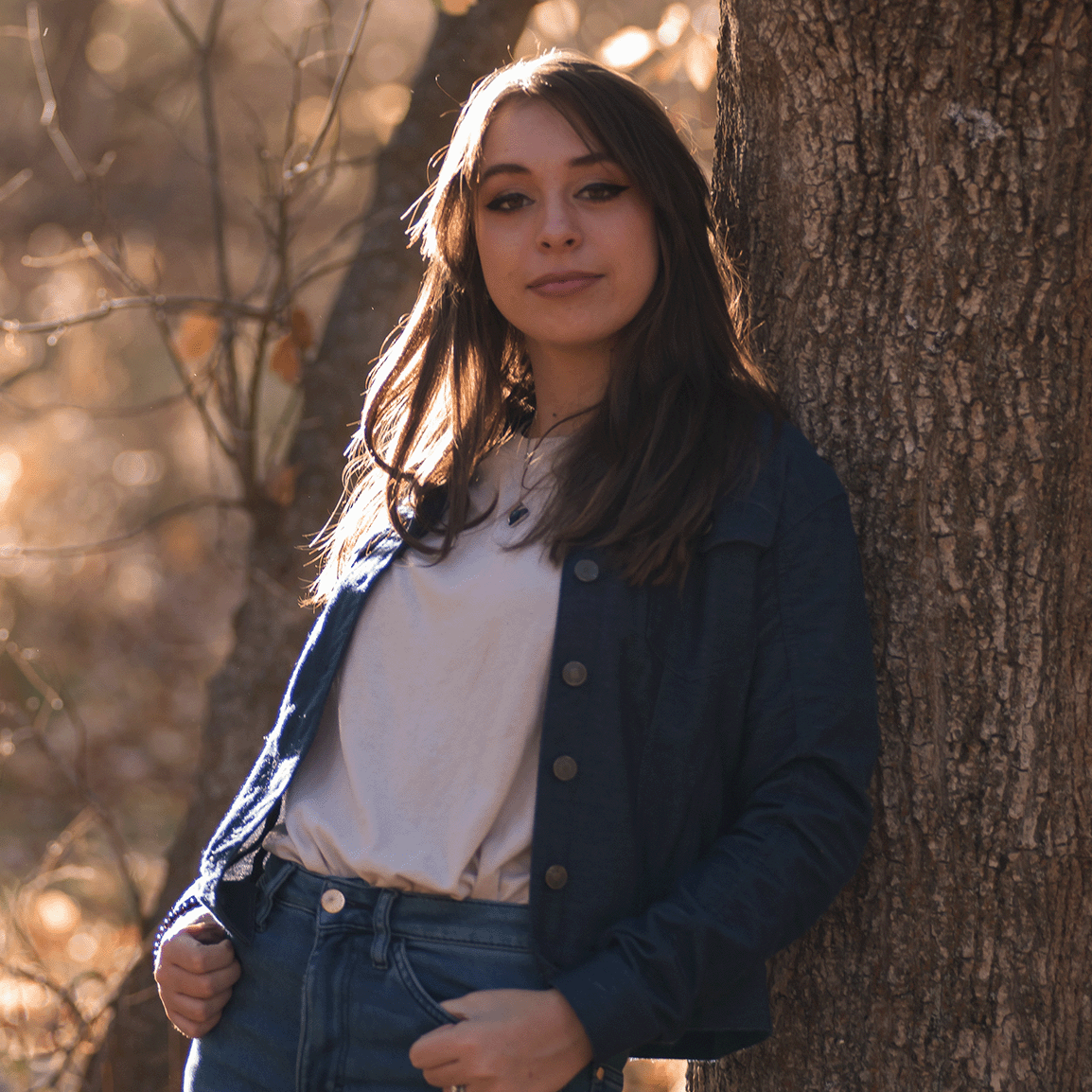This image shows Krista leaning against a tree. She has long dark hair and is dressed in a jean flannel and a white sweater. She is smiling at the camera