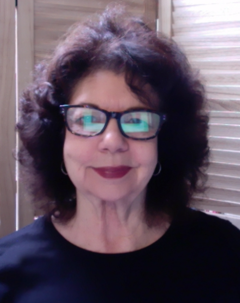 The image shows Mandi smiling at the camera with a maroon lipstick. She has a black shirt on and has short black hair and dark framed glasses. 
