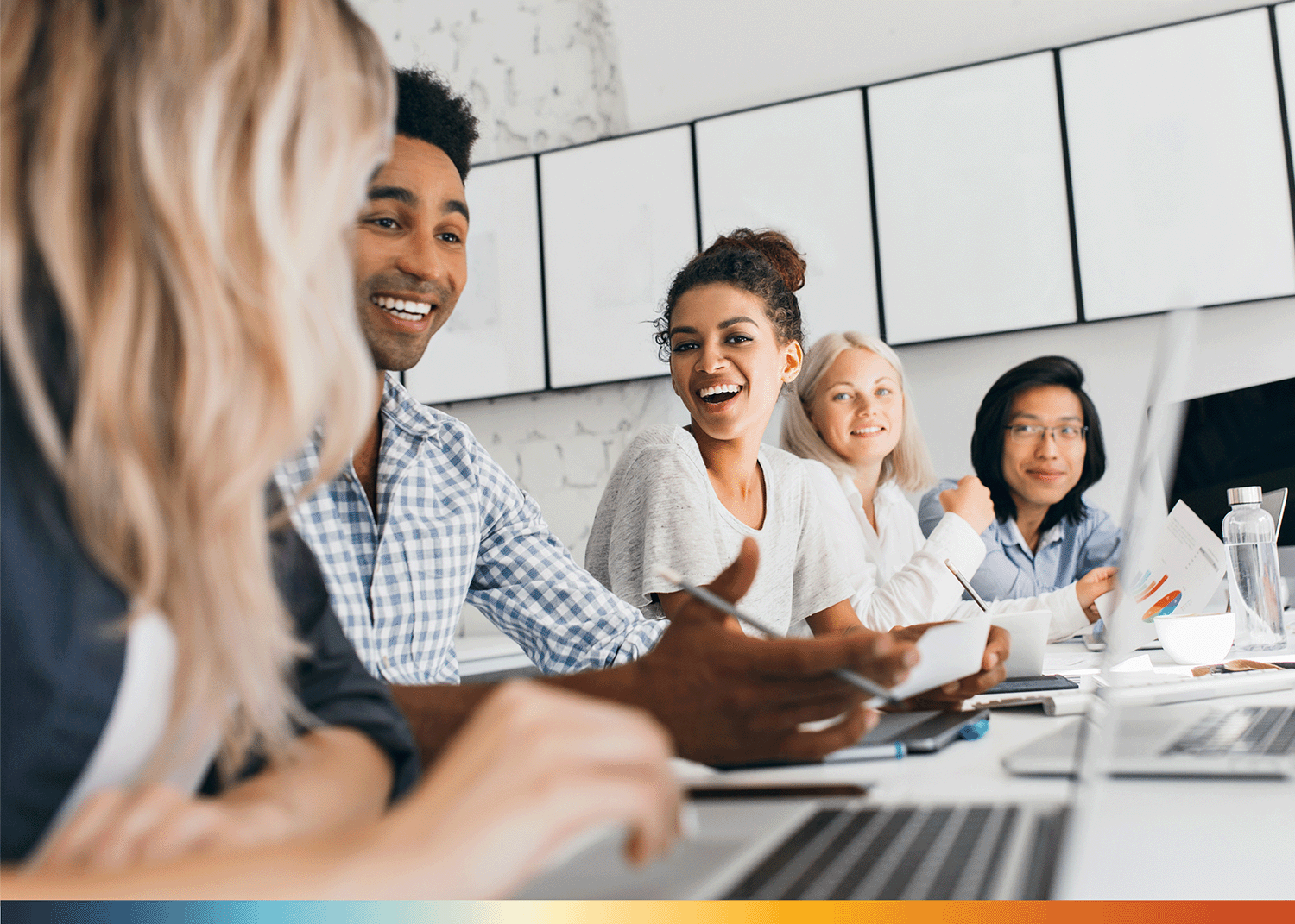 The Image portrays five people sitting in an office workplace. They are smiling and laughing. The woman in the center with curly hair is smiling directly towards the camera. At the bottom of the image is a gradient from blue to yellow to orange to red. 