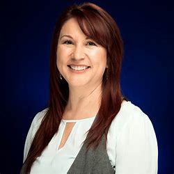 The image shows Sarai in a professional photo with a dark blue backdrop. She has long dark hair and is smiling brightly at the camera. She is wearing a white blouse with a grey vest.