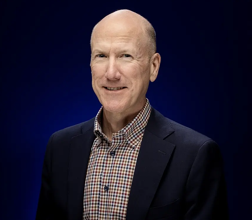The image shows Brian Heisterkamp smiling in a professional picture with a dark blue photos. He is wearing a dark suit with a plaid shirt. He is smiling at the camera