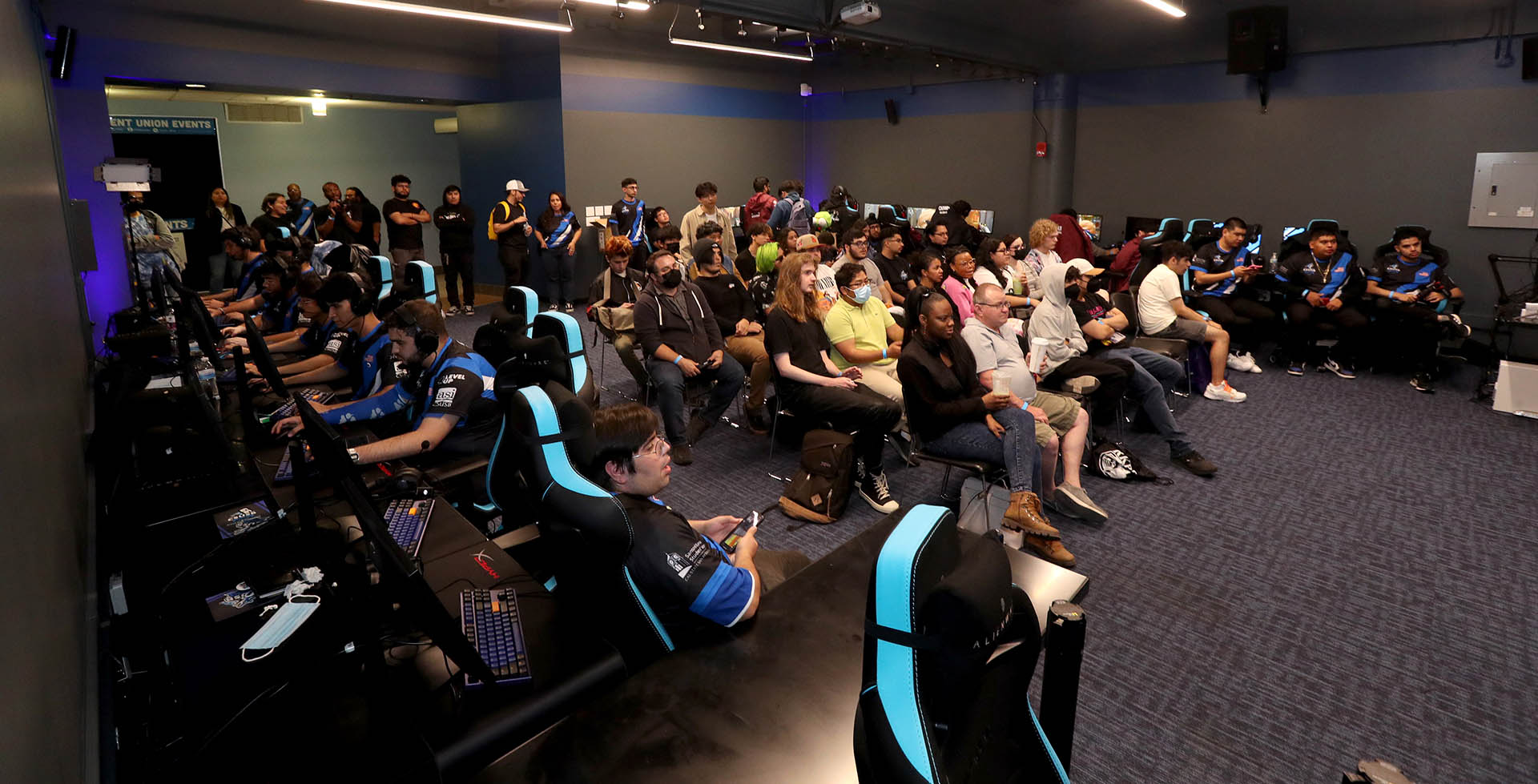 The audience watches on a big screen at the Esports Arena as the CSUSB Esports Club (left, on the computer keyboards) competes on a match.