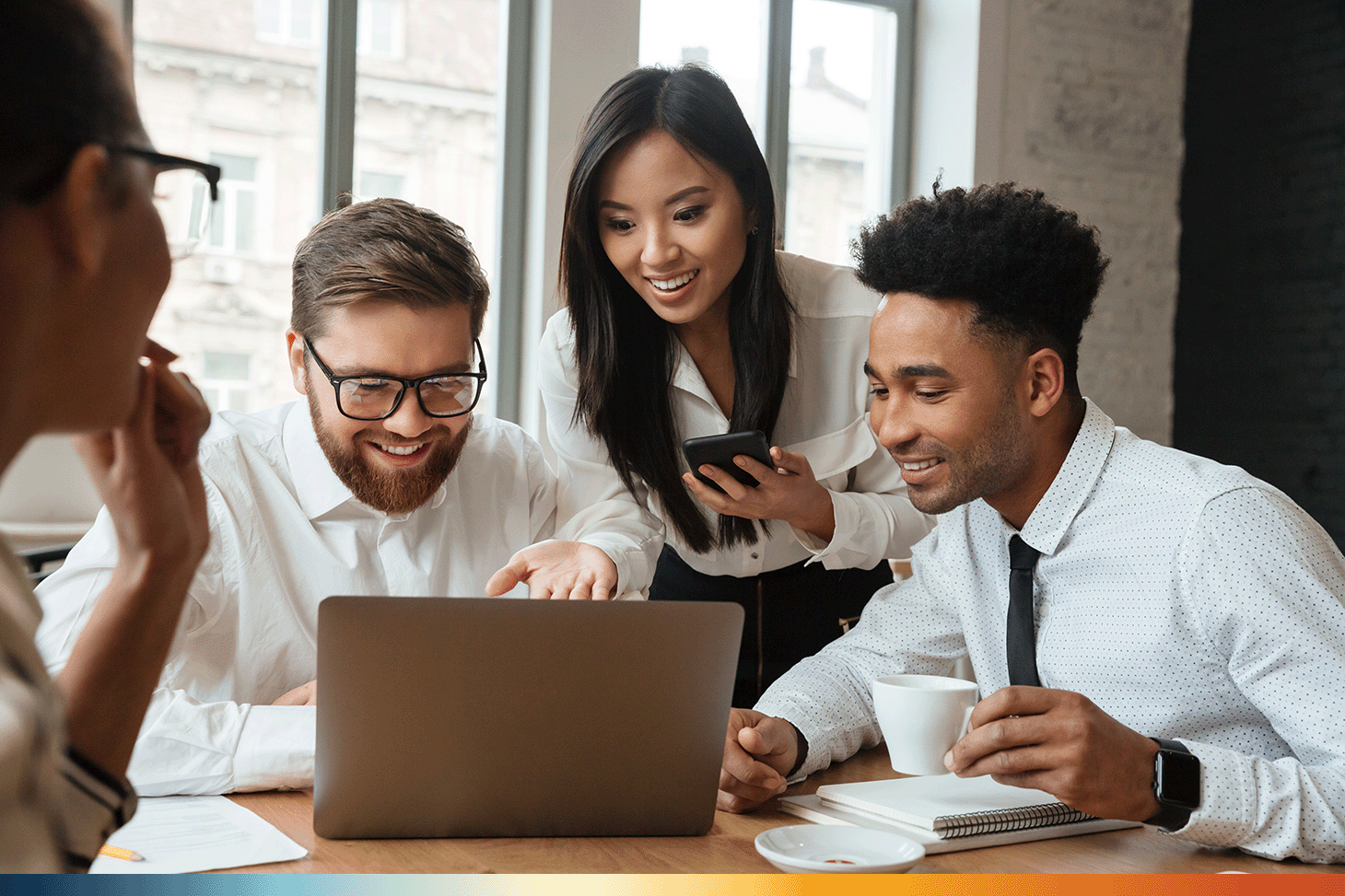 The Image shows 4 people sitting around a table all looking at a computer. Three people are facing the camera and one is faced away. They are all smiling at the computer and working together