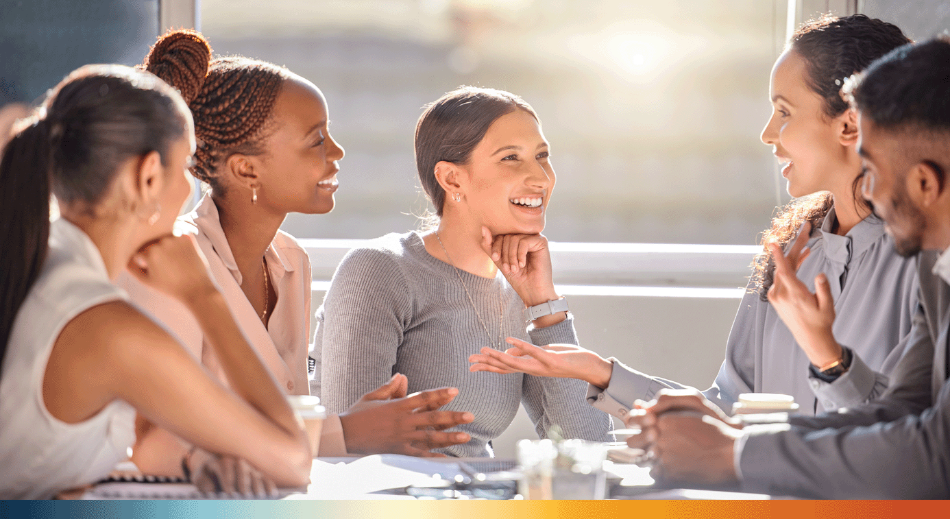 The Image shows 5 professional looking people sitting around a table smiling and talking to one another as they work together. 