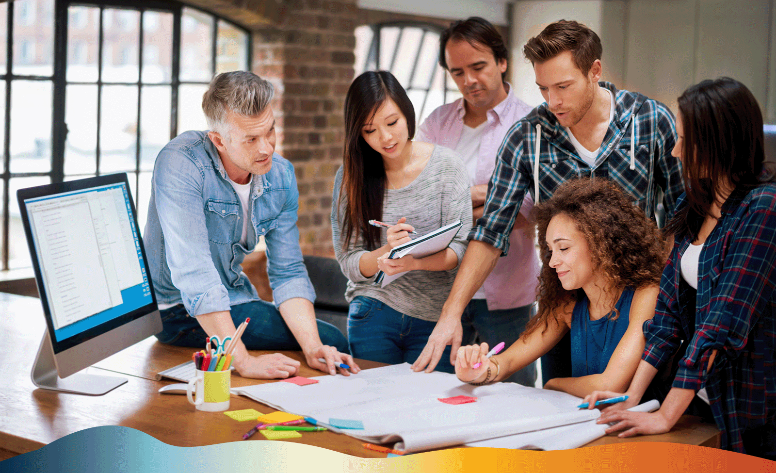 The image shows 6 people sitting and standing around a table working together as one man faces towards them all teaching them