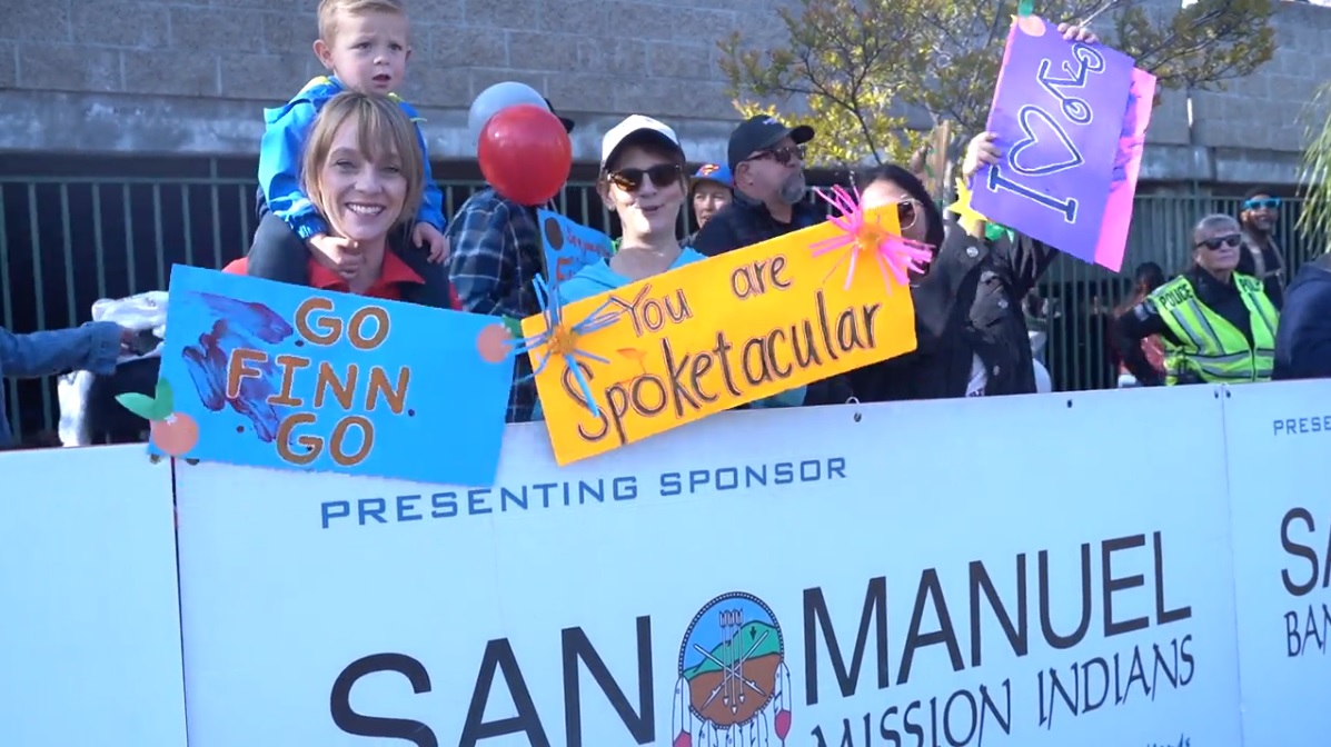 The image shows a crowd of people holding signs like, go Finn go, you are spoketacular, and I heart..., they look like they are supporters for people at the bike race