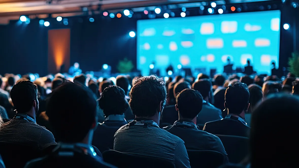 Photo of an audience at a conference, facing a stage.