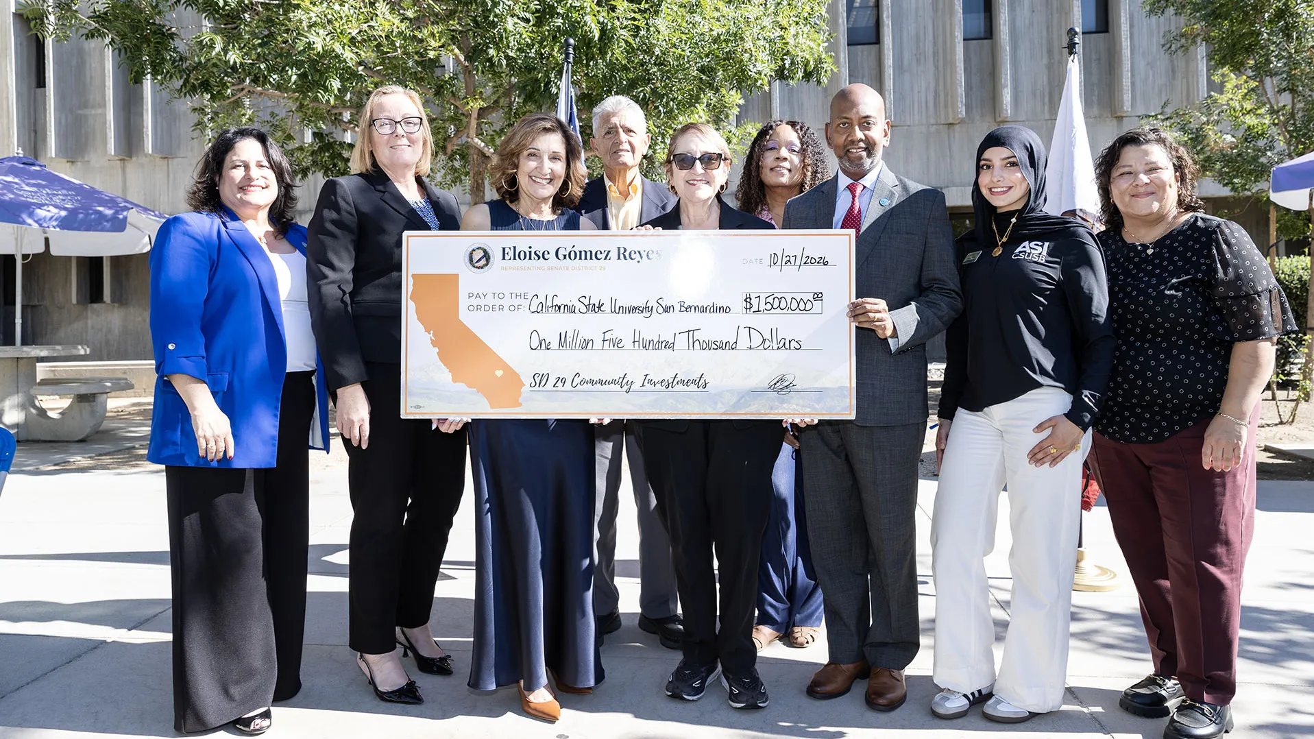 A group of nine community and university leaders smile while standing outside together and holding a large, ceremonial check for $1,500,000. The check is presented by Eloise Gómez Reyes for Senate District 29 Community Investments and is made out to California State University, San Bernardino.