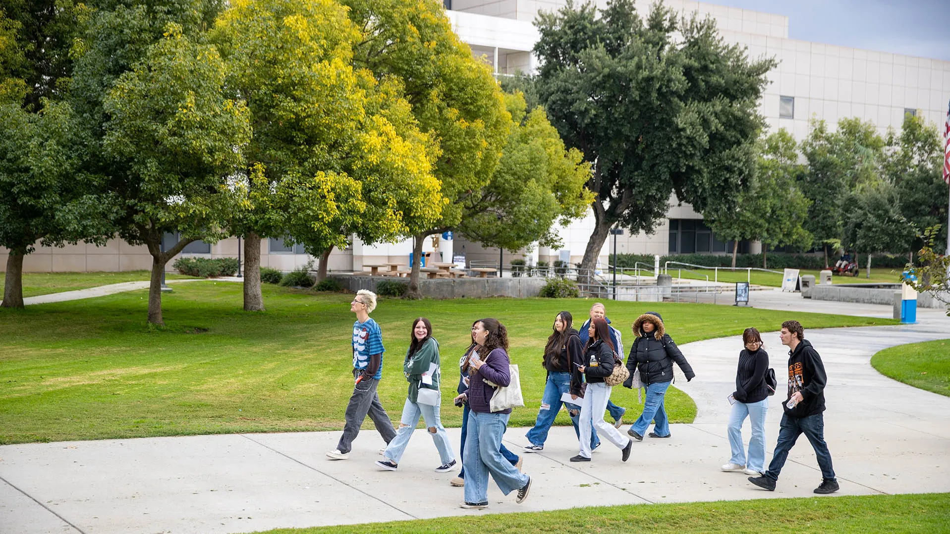 A group of diverse college students walking and chatting together along a concrete pathway, bordered by a grassy lawn on both sides, on the CSUSB campus with large trees and university buildings in the background.