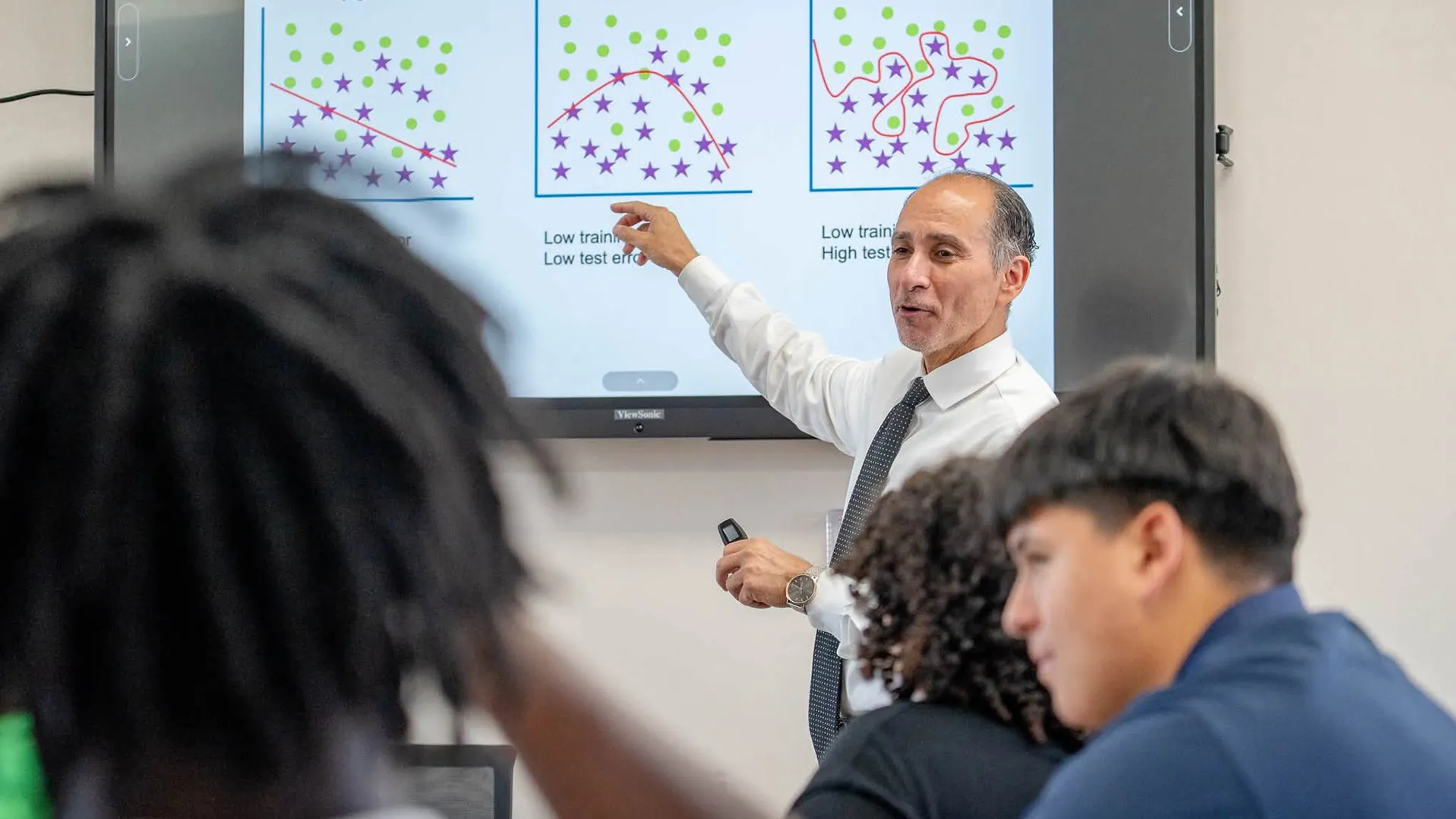 Nabeel Alzahrani, a CSUSB computer science faculty member, stands in front of a white board gesturing to writing on the board. Some people are in the foreground in a classroom setting.