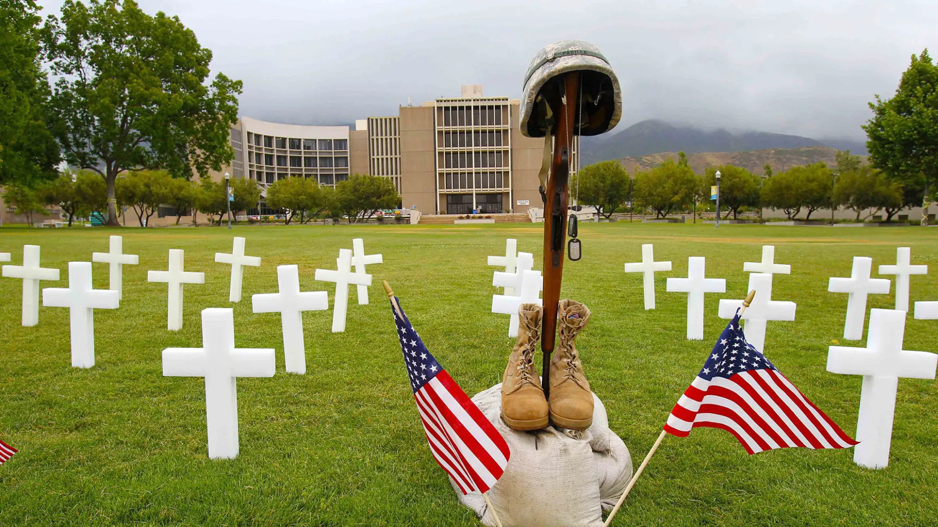 Photo of white crosses, military boots, rifle and helmet, two small American flags on the law in front of the Pfau Library.