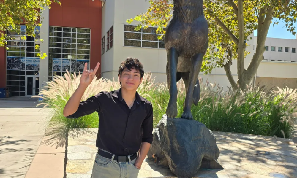 A male student in a black shirt and khaki pants holds a Coyote hand gesture beside a bronze howling coyote statue on a sunny, tree-lined campus.