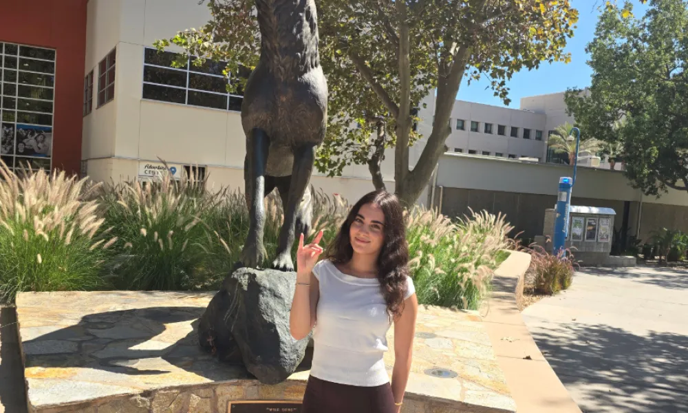 A female student in a white shirt and brown pants holds a Coyote hand gesture beside a bronze howling coyote statue on a sunny, tree-lined campus.