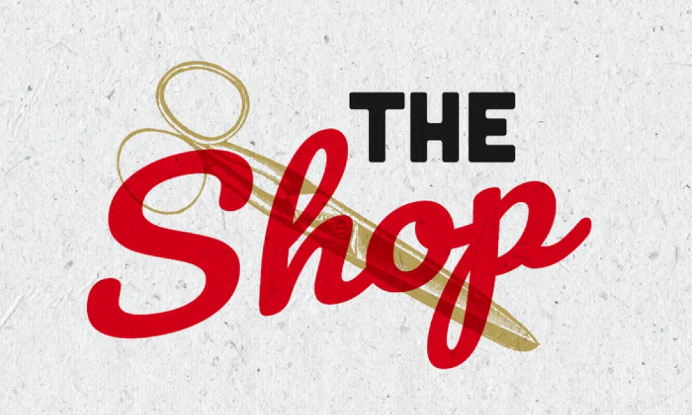 Logo with “THE” in small, bold black capital letters above “Shop” in large, red cursive script, on a light gray textured paper background. Behind the text is a gold line drawing of a pair of scissors.