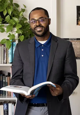 Marc Robinson, smiling and reading a book in front of a bookshelf