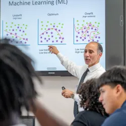 Nabeel Alzahrani, a CSUSB computer science faculty member, stands in front of a white board gesturing to writing on the board. Some people are in the foreground in a classroom setting.