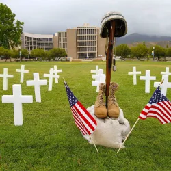 Photo of white crosses, military boots, rifle and helmet, two small American flags on the law in front of the Pfau Library.