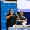Two presenters stand at the front of a California State University, San Bernardino (CSUSB) event. The person on the left speaks into a handheld microphone while gesturing with one hand, and the person on the right holds papers and looks toward the speaker. A table with promotional items is in the foreground, and a blue CSUSB-branded backdrop and presentation screen are behind them.