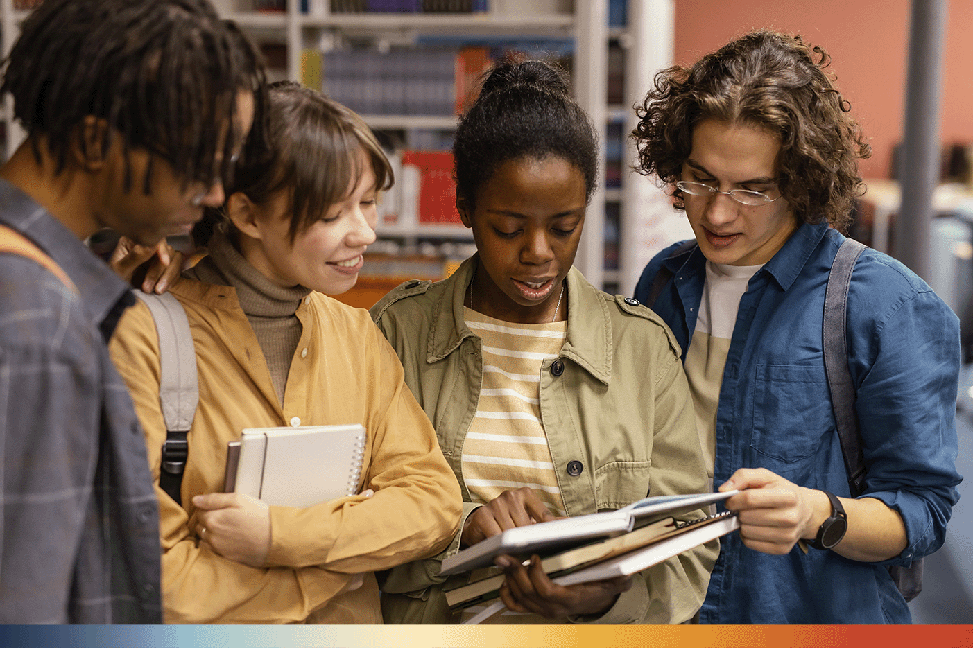 The Image shows 4 students smiling and huddled closer together faced towards the camera. They are all looking at the notebooks of the girl in the middle. 