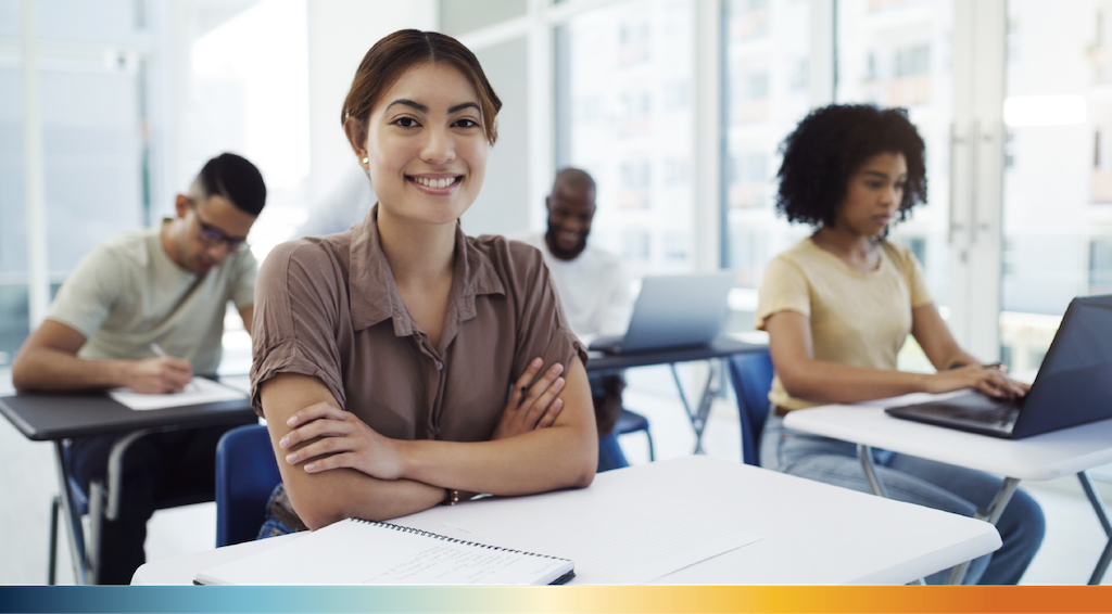 A student sitting at a desk and smiling at the camera in a classroom with other students in the background.