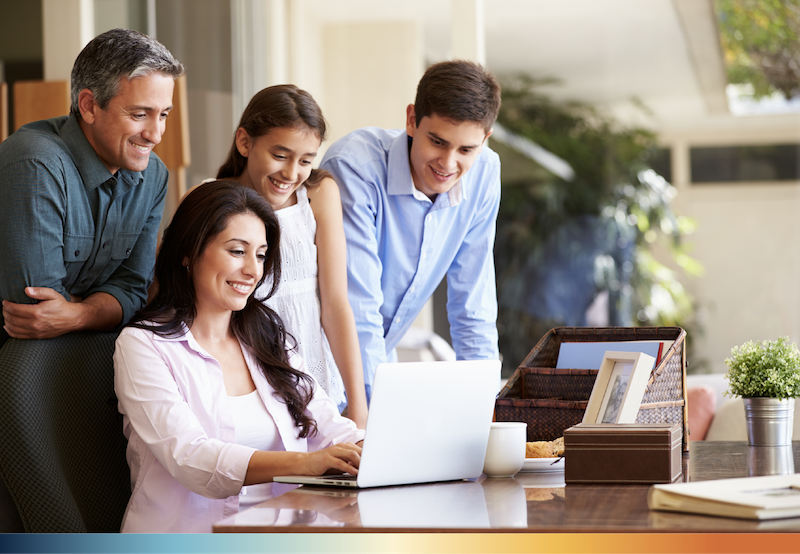 A family of four gathered at a desk and look at a laptop together.
