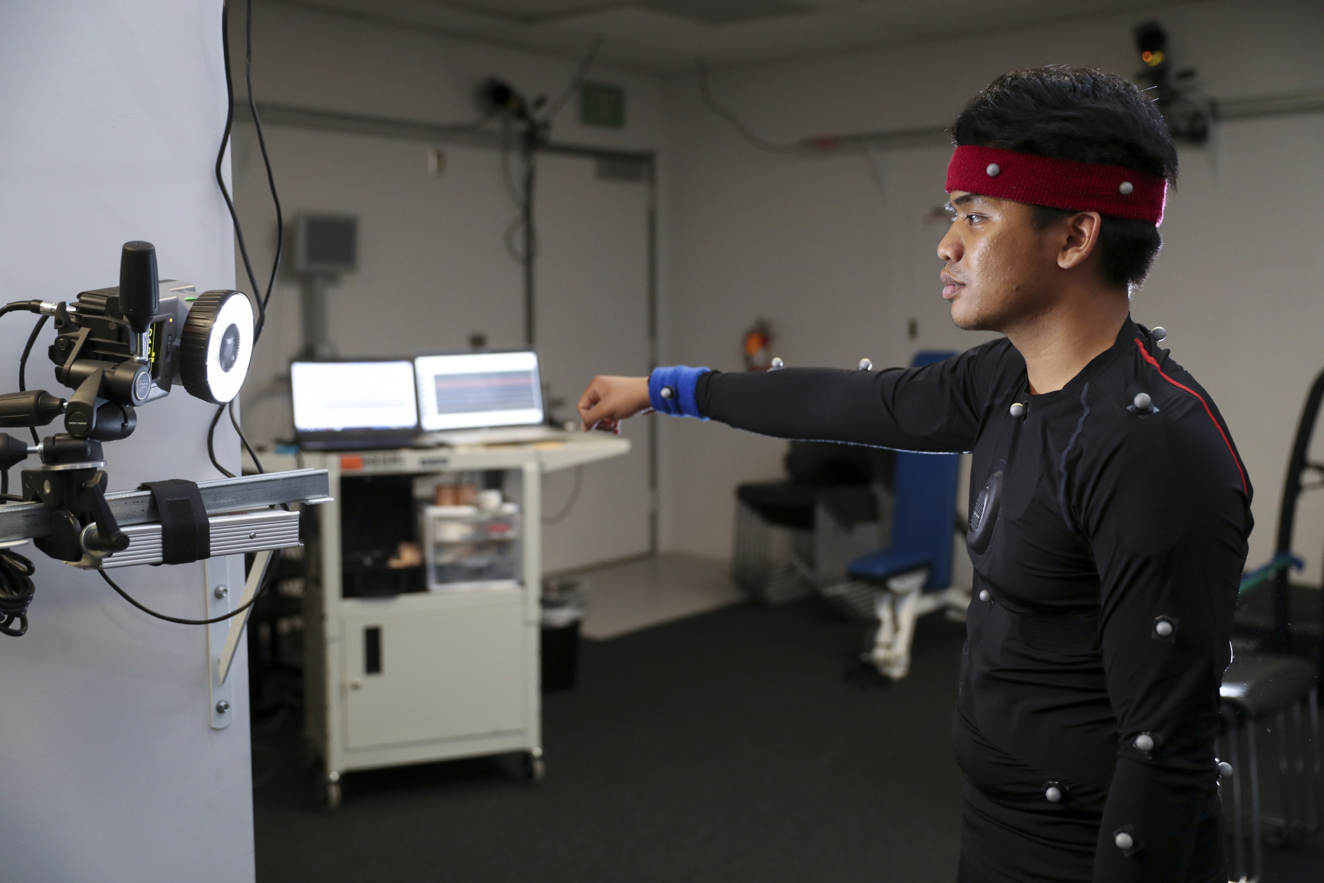 A person wearing a motion capture suit stands in a lab, extending one arm forward toward a mounted camera. Reflective motion capture markers are attached to their suit and headband, while computers and lab equipment are visible in the background.