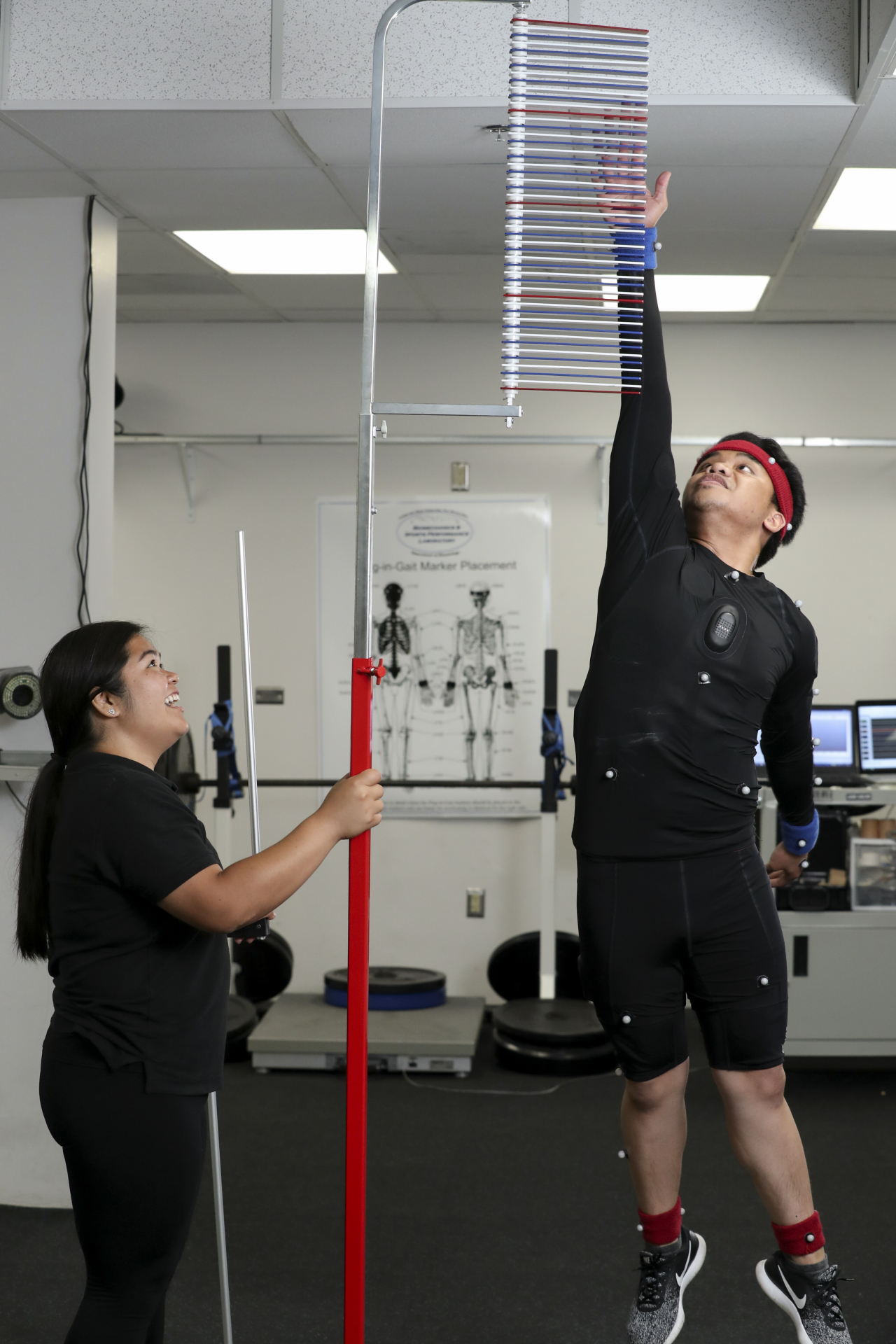 A person wearing a motion capture suit and headband reaches upward to touch measurement vanes on a vertical jump testing device, while another person beside them holds the pole steady and smiles. The scene takes place in a motion capture lab with fitness equipment and computer monitors in the background.