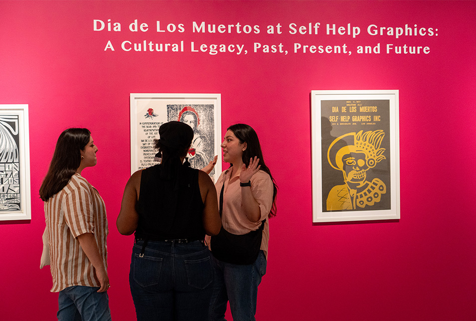 A group of three women talk in front of a wall with mounted artwork