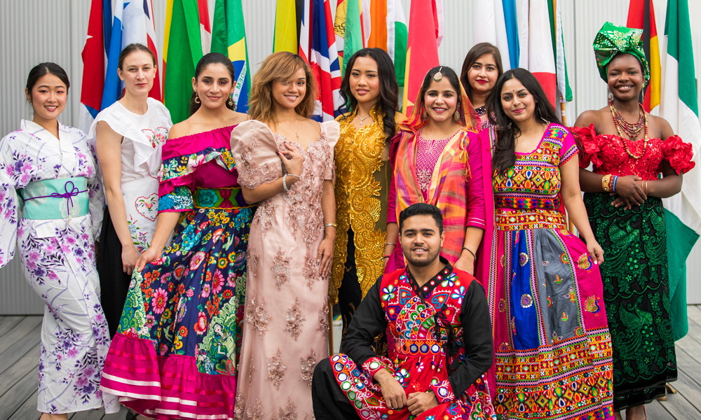 A group of international students dressed in colorful traditional clothing from different countries stand smiling in front of global flags, celebrating cultural diversity and international education.