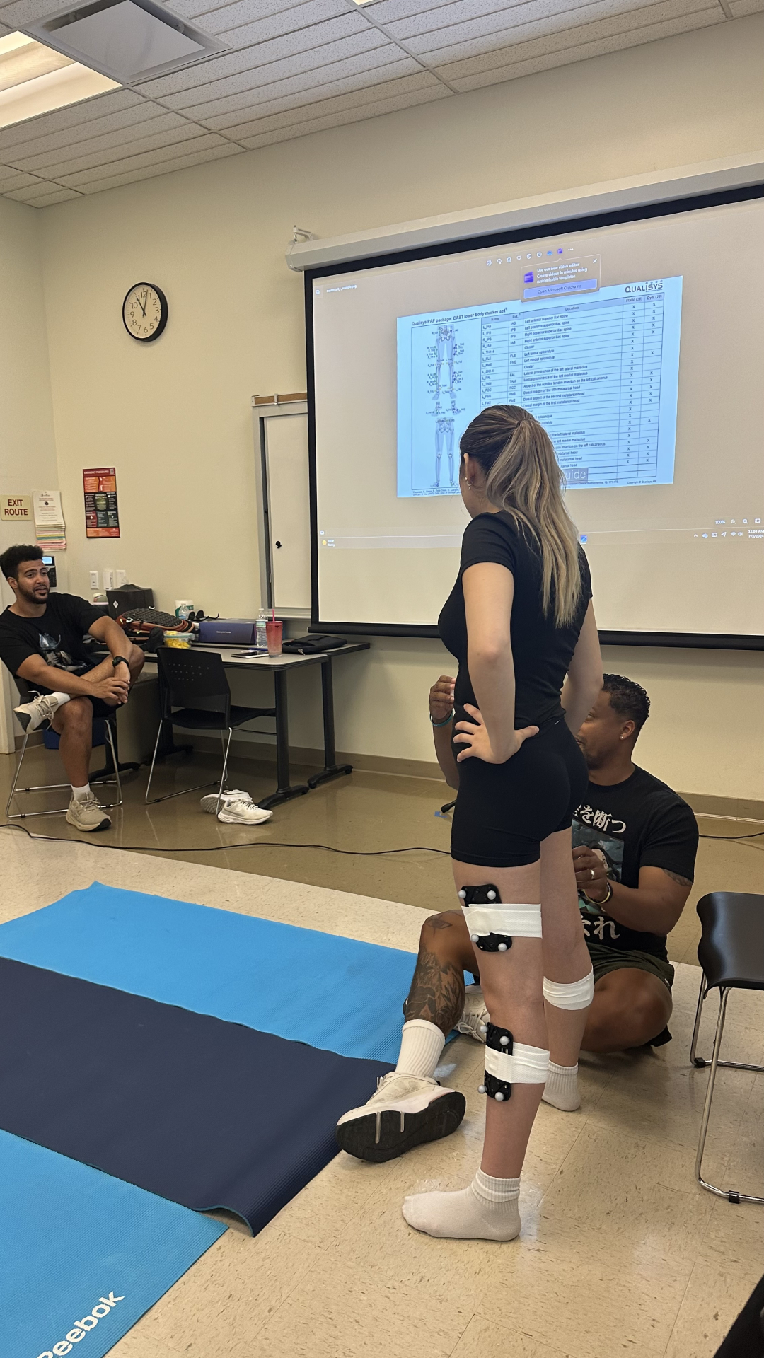 A student wearing motion capture sensors on her legs stands in front of a classroom projector screen displaying a Qualisys marker placement chart, while an instructor kneels nearby adjusting equipment and classmates observe the demonstration.