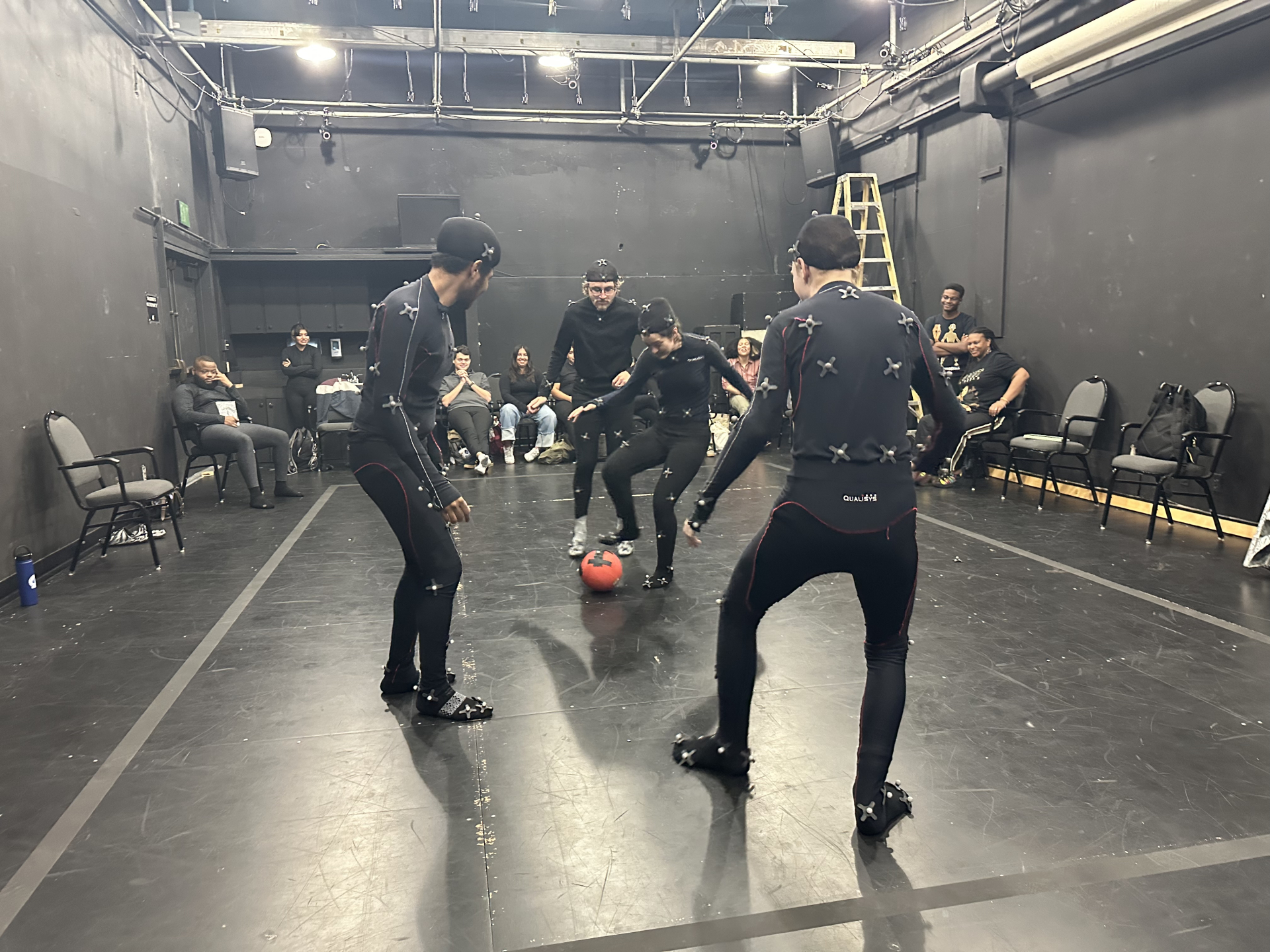 Four students wearing full-body motion capture suits play soccer with a red ball inside CSUSB’s Motion Capture Studio, while classmates and instructors observe from the sides in the black-walled lab space.