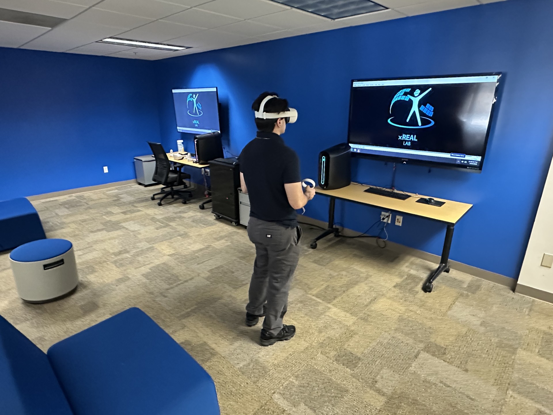 Alt text: A person wearing a VR headset and holding controllers stands in a blue-walled lab space. A large screen on the wall displays the xREAL Lab logo, mirrored from the headset. Nearby, a computer workstation with a chair, monitor, and equipment cart is set up against the wall. The room has beige carpet and blue lounge seating.