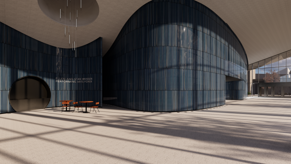 Interior view of the Ellen and Stan Weisser Performing Arts Foyer. The space features a modern, curved blue wall with vertical paneling, a round entrance on the left, minimalist furniture with orange chairs, and large floor-to-ceiling windows casting shadows across the floor.