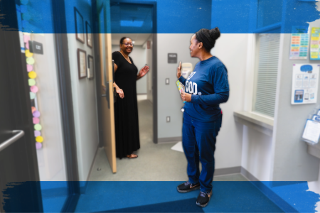 Two women stand in a hallway beside a blue sign, engaged in conversation.