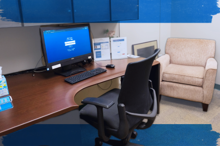 Office setting with a wooden desk, computer showing a login screen, black chair, hand sanitizer, framed pictures, and cozy beige armchair, creating a professional atmosphere.