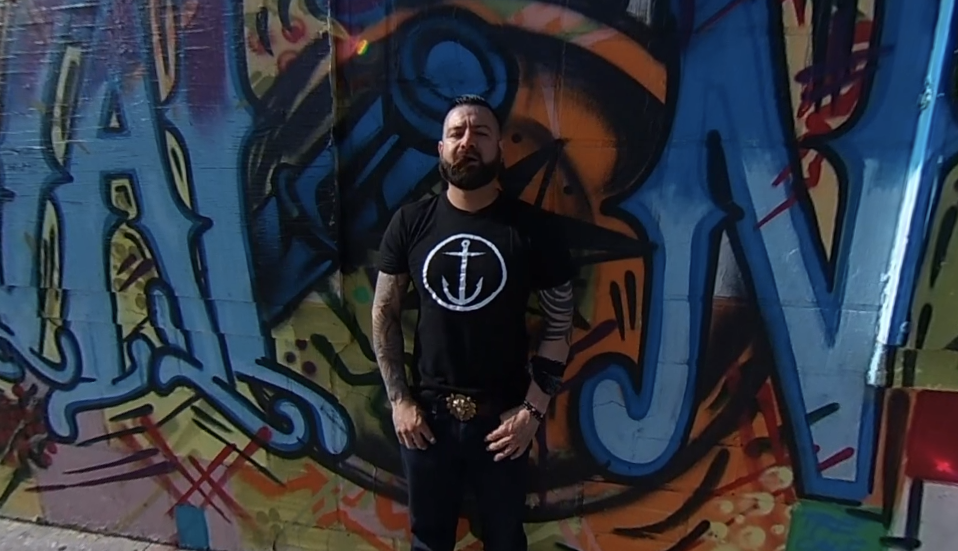 **Alt Text:** A man stands confidently in front of a colorful graffiti-covered wall. He is wearing a black T-shirt with a white anchor symbol, dark jeans, and a decorative belt buckle. His arms are tattooed, and he has a beard and styled hair. The vivid graffiti behind him features bold blue, orange, and yellow patterns, creating a striking urban backdrop.