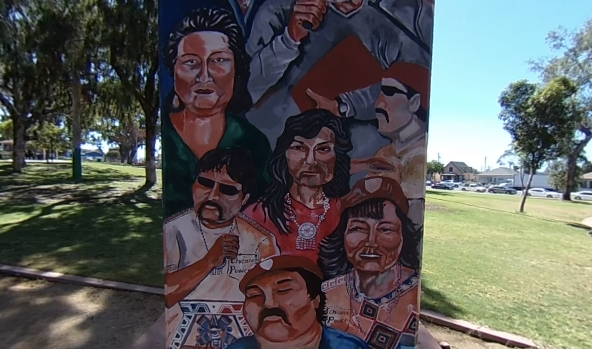 **Alt Text:** A colorful mural painted on a concrete pillar depicts several people in a bold, expressive style. The figures wear clothing with cultural and political significance, including shirts and hats labeled “Chicana Power.” The mural is set in an outdoor park with trees, grass, and parked cars visible in the background, celebrating community pride and heritage.