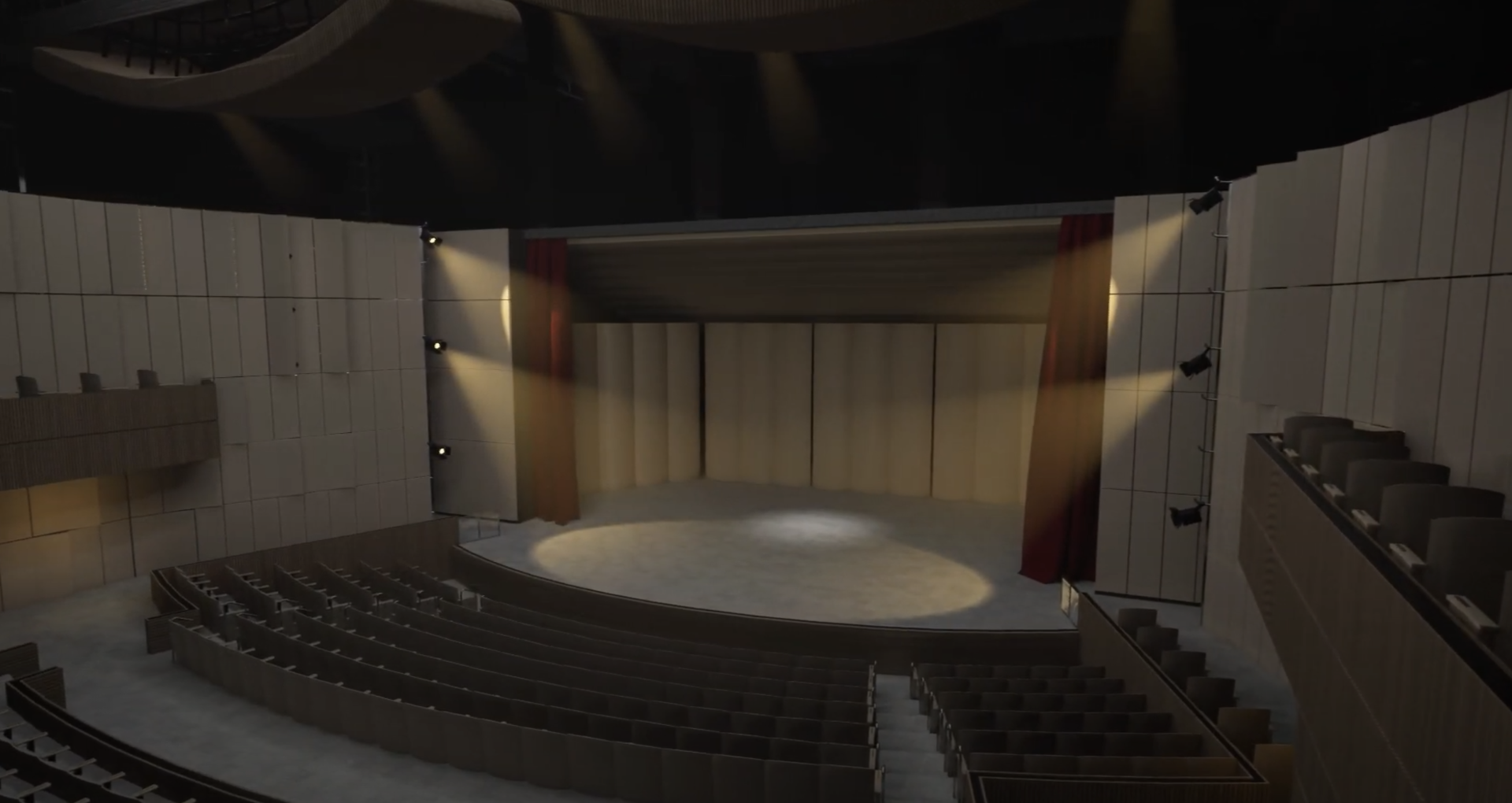 **Alt Text:** A wide-angle view of an empty theater auditorium with rows of curved seating facing a stage. The stage is illuminated by several spotlights, with red curtains framing the sides and a light-colored backdrop behind. The modern interior features wooden acoustic panels and overhead lighting fixtures, creating a warm and professional atmosphere suitable for performances or presentations.