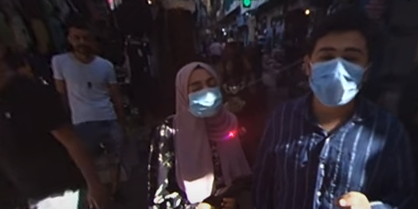 Here’s an appropriate alt text description for that image: **Alt text:** Two people wearing face masks walk through a crowded outdoor market. The person on the left wears a pink headscarf and dark clothing, while the person on the right wears a blue striped shirt. The market is busy with other people and lined with shops and hanging goods in the background.