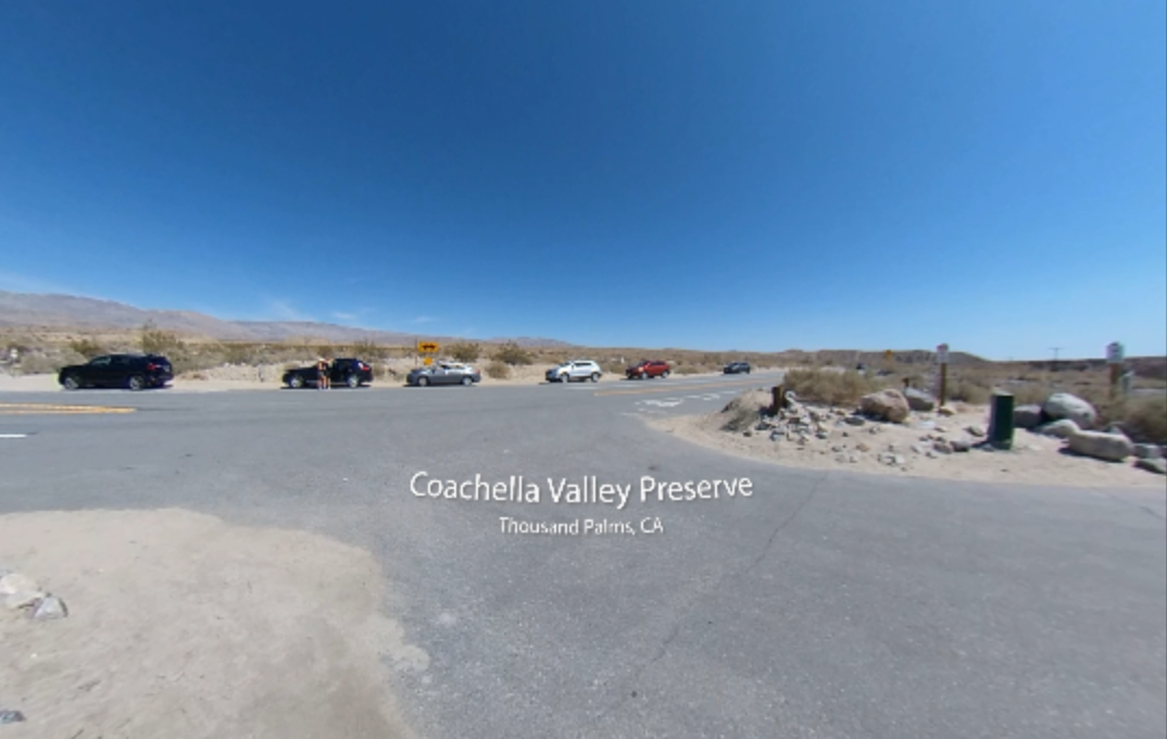 A 360-degree image of the Coachella Valley Preserve entrance in Thousand Palms, California. The foreground shows a sandy and paved road intersection with several parked cars. Desert landscape, scattered boulders, and dry brush stretch across the background, with distant mountains under a bright blue sky. The text “Coachella Valley Preserve, Thousand Palms, CA” is centered at the bottom of the image.