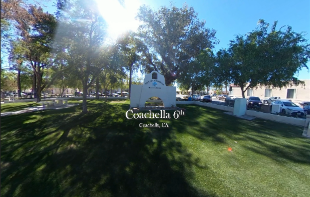 A 360-degree image of a grassy park area in Coachella, California. In the center is a white mission-style city landmark structure labeled “Coachella 6th.” Tall trees create shaded areas while sunlight filters through the branches. Park benches and nearby streets with parked cars surround the scene. The text “Coachella 6th, Coachella, CA” appears in the middle of the image.