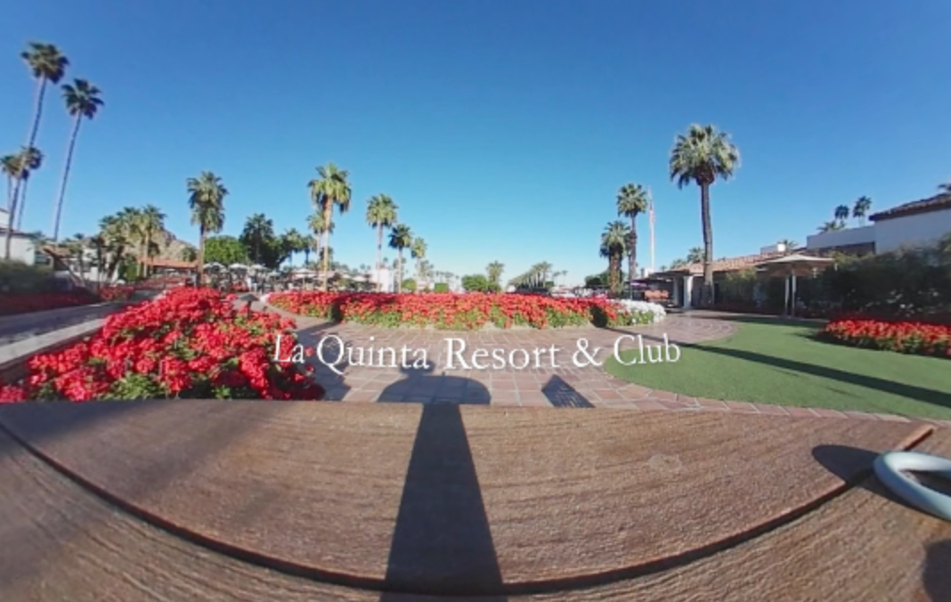 A 360-degree image showing the entrance area of La Quinta Resort & Club. The view includes vibrant red flowers arranged in garden beds, palm trees lining the background, and tiled walkways. The text “La Quinta Resort & Club” is centered in the image. Clear blue skies and resort buildings frame the scenic desert landscape.