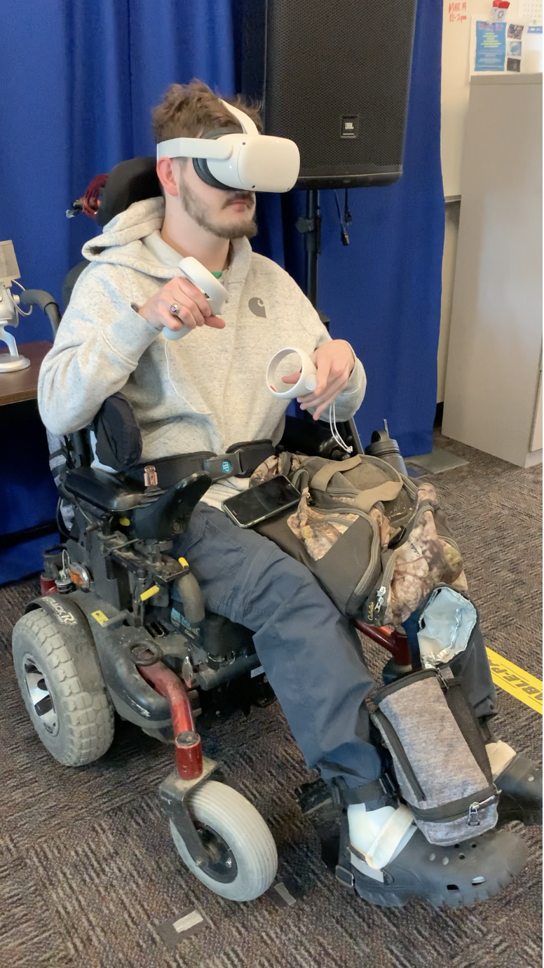 A young man seated in a motorized wheelchair is using a virtual reality headset and handheld VR controllers. He is wearing a gray hoodie and dark pants, with personal items such as a camouflage bag and smartphone resting on his lap. The background includes a blue curtain, a speaker on a stand, and part of a bulletin board and cabinet, suggesting an indoor media or technology lab setting.