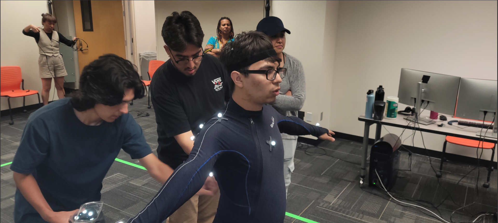 Students at CSUSB work together to calibrate a motion capture suit, adjusting reflective markers on a participant standing in a T-pose while instructors and classmates observe in the background inside the xREAL Lab’s Motion Capture Studio.