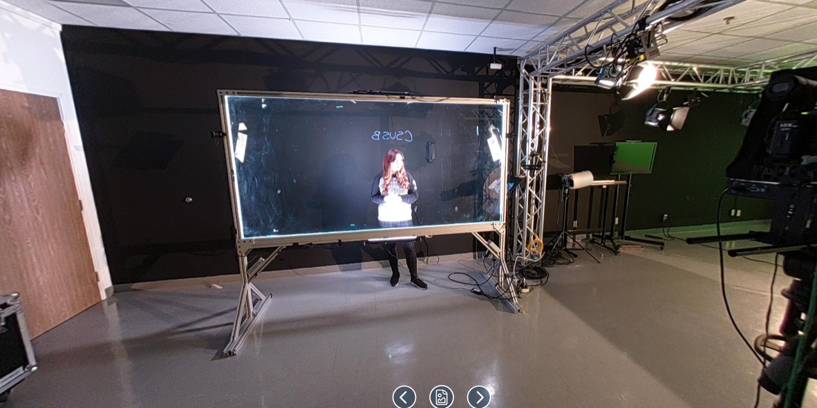 A studio setup featuring a lightboard recording session at California State University, San Bernardino. A woman stands behind a large transparent glass panel, writing on it with a marker while facing the camera, which captures the writing in a mirrored format for viewers. The word "CSUSB" is visible in reverse on the board. The studio is equipped with professional lighting mounted on a truss system, and a camera is set up in the foreground. The walls are painted dark to reduce reflections, and additional production equipment is arranged along the sides of the room.