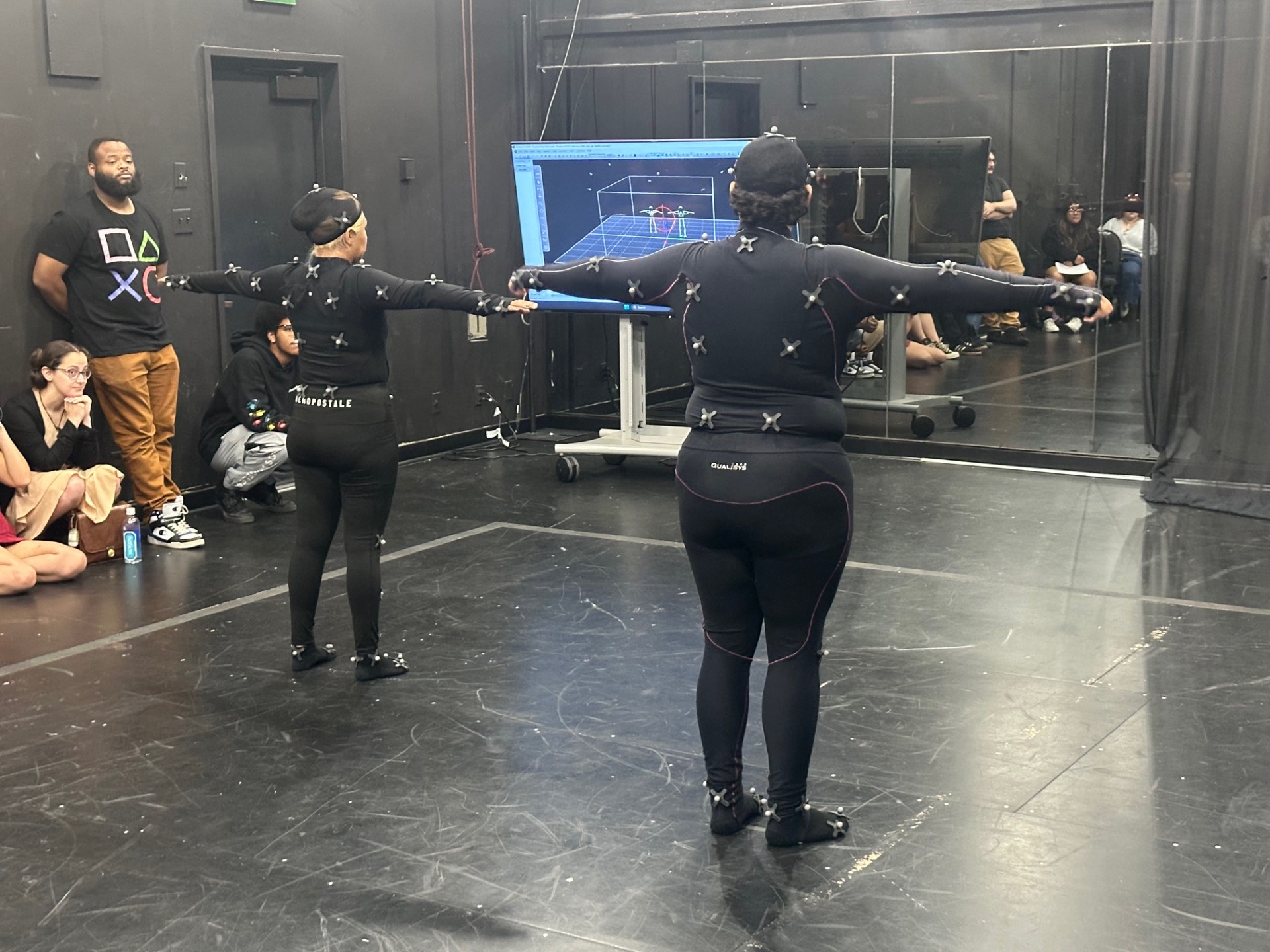 Two individuals wearing full motion capture suits stand side by side in a T-pose in a studio space. A group of students sits on the floor and stands against the walls observing. A large screen displays live tracking data of the performers’ skeletal motion capture.